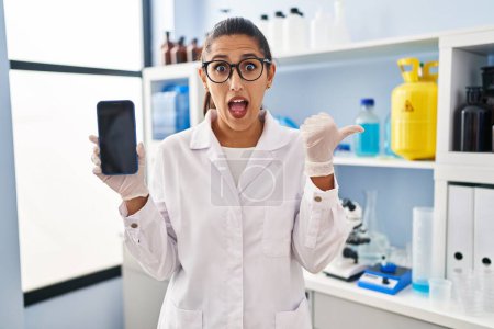 Foto de Joven mujer hispana trabajando en laboratorio científico con smartphone apuntando con el pulgar hacia un lado sonriendo feliz con la boca abierta - Imagen libre de derechos
