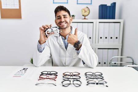 Photo for Young optician man holding optometry glasses doing happy thumbs up gesture with hand. approving expression looking at the camera showing success. - Royalty Free Image