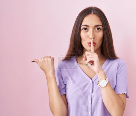 Photo for Young hispanic woman with long hair standing over pink background asking to be quiet with finger on lips pointing with hand to the side. silence and secret concept. - Royalty Free Image