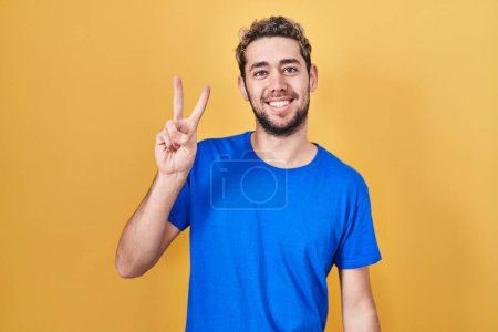 Téléchargez les photos : Homme hispanique avec la barbe debout sur fond jaune souriant avec le visage heureux clin d'oeil à la caméra faisant signe de victoire avec les doigts. numéro deux. - en image libre de droit