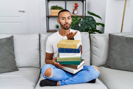 Photo for African american young man holding a pile of books sitting on the sofa looking at the camera blowing a kiss with hand on air being lovely and sexy. love expression. - Royalty Free Image
