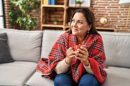 Photo for Middle age hispanic woman drinking coffee covering with blanket at home - Royalty Free Image