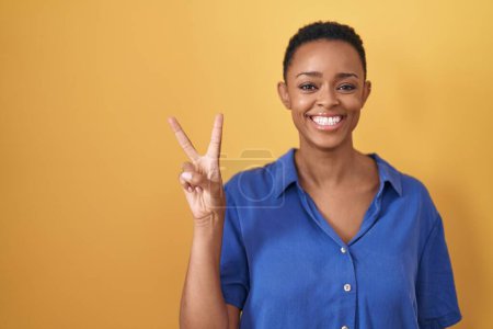 Foto de Mujer afroamericana de pie sobre fondo amarillo mostrando y señalando hacia arriba con los dedos número dos mientras sonríe confiado y feliz. - Imagen libre de derechos