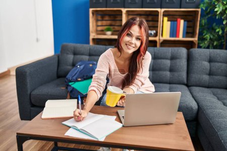 Photo for Young caucasian woman student sitting on sofa writing on notebook drinking coffee at home - Royalty Free Image