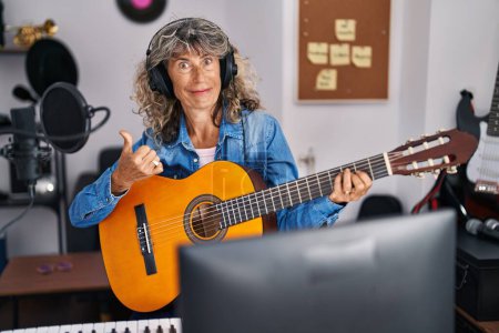 Téléchargez les photos : Femme du moyen âge jouant de la guitare classique au studio de musique pointant le pouce vers le côté souriant heureux avec la bouche ouverte - en image libre de droit