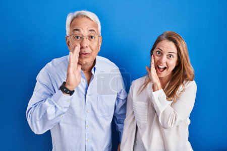 Téléchargez les photos : Couple hispanique du Moyen Age debout sur fond bleu main sur bouche racontant rumeur secrète, chuchotant conversation malicieuse - en image libre de droit