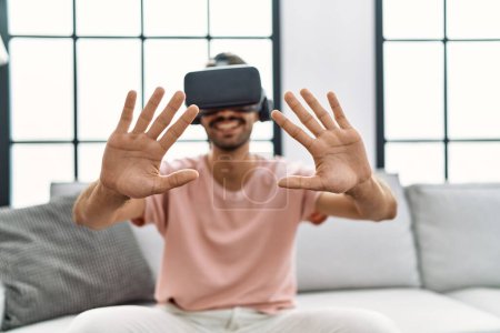 Téléchargez les photos : Bel homme hispanique portant des lunettes de réalité virtuelle au salon à la maison - en image libre de droit