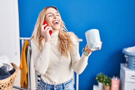 Photo for Young blonde woman talking on smartphone drinking coffee waiting for washing machine at laundry room - Royalty Free Image