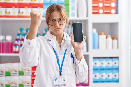 Foto de Young caucasian woman working at pharmacy drugstore showing smartphone screen annoyed and frustrated shouting with anger, yelling crazy with anger and hand raised - Imagen libre de derechos