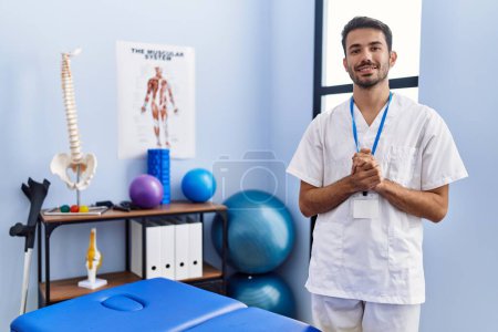 Foto de Young hispanic man wearing physiotherapist uniform standing at rehab clinic - Imagen libre de derechos