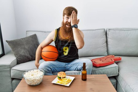 Photo for Caucasian man with long beard holding basketball ball cheering tv game covering one eye with hand, confident smile on face and surprise emotion. - Royalty Free Image