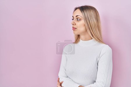 Photo for Young blonde woman wearing white sweater over pink background looking to side, relax profile pose with natural face with confident smile. - Royalty Free Image