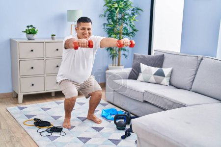 Photo for Young latin man training leg exercise using dumbbells at home - Royalty Free Image