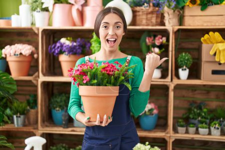 Photo for Young hispanic woman working at florist shop holding plant pointing thumb up to the side smiling happy with open mouth - Royalty Free Image