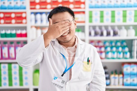 Téléchargez les photos : Jeune homme chinois travaillant à la pharmacie pharmacie souriant et riant avec la main sur le visage couvrant les yeux pour la surprise. concept aveugle. - en image libre de droit