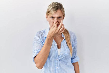 Photo for Beautiful blonde woman standing over white background smelling something stinky and disgusting, intolerable smell, holding breath with fingers on nose. bad smell - Royalty Free Image