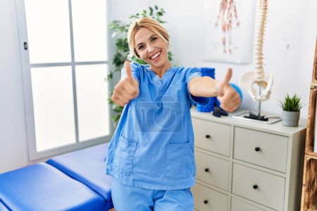 Photo for Beautiful blonde physiotherapist woman working at pain recovery clinic approving doing positive gesture with hand, thumbs up smiling and happy for success. winner gesture. - Royalty Free Image