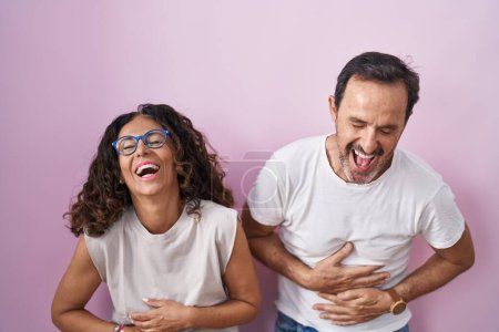 Téléchargez les photos : Couple hispanique du moyen âge ensemble sur fond rose souriant et riant fort à haute voix parce que drôle blague folle avec les mains sur le corps. - en image libre de droit