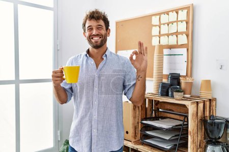 Foto de Joven hombre guapo bebiendo una taza de café en la oficina haciendo ok signo con los dedos, sonriendo gesto amistoso excelente símbolo - Imagen libre de derechos