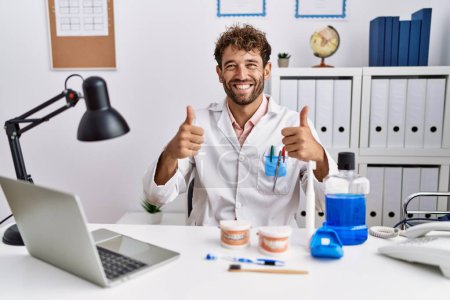 Young hispanic dentist man working at medical clinic success sign doing positive gesture with hand, thumbs up smiling and happy. cheerful expression and winner gesture. 