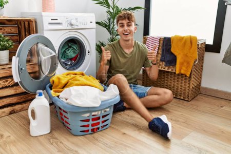 Foto de Young caucasian man putting dirty laundry into washing machine success sign doing positive gesture with hand, thumbs up smiling and happy. cheerful expression and winner gesture. - Imagen libre de derechos