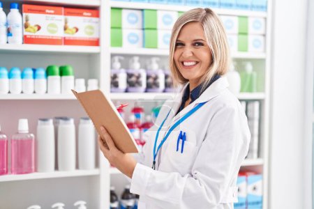 Photo for Young blonde woman pharmacist smiling confident reading notebook at pharmacy - Royalty Free Image