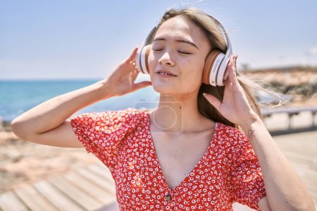 Foto de Joven chica china sonriendo feliz escuchando música en la playa. - Imagen libre de derechos