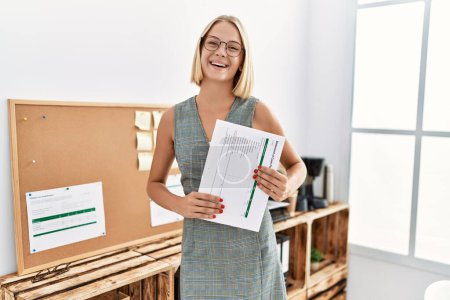 Foto de Joven mujer rubia sonriendo confiado sosteniendo documento en la oficina - Imagen libre de derechos
