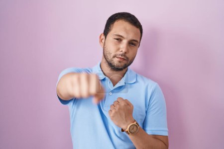 Photo for Hispanic man standing over pink background punching fist to fight, aggressive and angry attack, threat and violence - Royalty Free Image