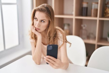 Téléchargez les photos : Jeune femme blonde utilisant un smartphone avec une expression sérieuse à la maison - en image libre de droit