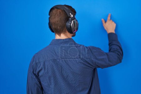 Photo for Hispanic man with beard listening to music wearing headphones posing backwards pointing ahead with finger hand - Royalty Free Image