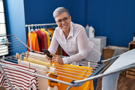 Téléchargez les photos : Middle age woman using smartphone hanging clothes on clothesline at laundry room - en image libre de droit