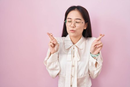 Téléchargez les photos : Jeune femme chinoise debout sur fond rose geste doigt croisé souriant avec espoir et les yeux fermés. chance et concept superstitieux. - en image libre de droit