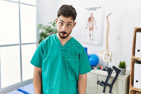 Photo for Young man with beard working at pain recovery clinic making fish face with lips, crazy and comical gesture. funny expression. - Royalty Free Image