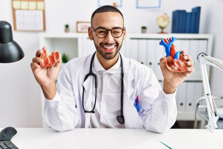 Téléchargez les photos : Afro-américain médecin homme tenant coeur anatomique à la clinique clin d'oeil en regardant la caméra avec expression sexy, gai et heureux visage. - en image libre de droit