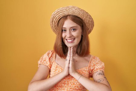 Foto de Joven pelirroja de pie sobre fondo amarillo con sombrero de verano rezando con las manos juntas pidiendo perdón sonriendo confiado. - Imagen libre de derechos