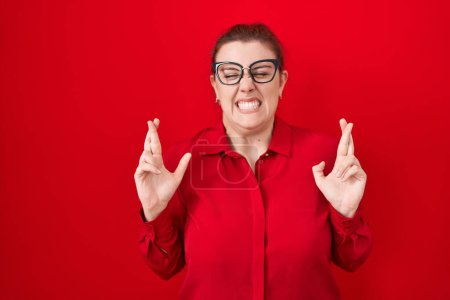 Foto de Young hispanic woman with red hair standing over red background gesturing finger crossed smiling with hope and eyes closed. luck and superstitious concept. - Imagen libre de derechos