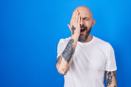 Photo for Hispanic man with tattoos standing over blue background yawning tired covering half face, eye and mouth with hand. face hurts in pain. - Royalty Free Image