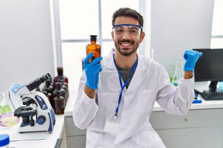 Photo for Young hispanic man working at scientist laboratory holding bottle pointing thumb up to the side smiling happy with open mouth - Royalty Free Image