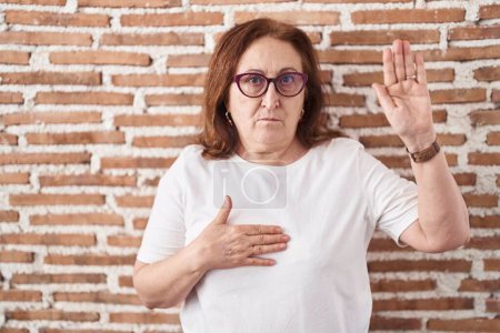 Foto de Mujer mayor con gafas de pie sobre ladrillos juramento de la pared con la mano en el pecho y la palma abierta, haciendo una promesa de lealtad juramento - Imagen libre de derechos
