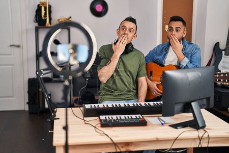 Foto de Dos hombres tocando guitarra clásica en el estudio de música grabando la canción cubriendo la boca con la mano, conmocionados y temerosos de equivocarse. expresión sorprendida - Imagen libre de derechos