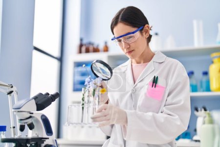 Foto de Mujer joven vistiendo uniforme científico buscando flores con lupa en el laboratorio - Imagen libre de derechos