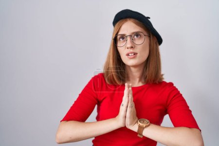 Foto de Young redhead woman standing wearing glasses and beret begging and praying with hands together with hope expression on face very emotional and worried. begging. - Imagen libre de derechos