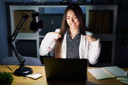 Photo for Young brunette woman working at the office at night with laptop looking confident with smile on face, pointing oneself with fingers proud and happy. - Royalty Free Image