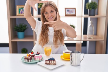 Téléchargez les photos : Jeune femme caucasienne mangeant des pâtisseries t pour le petit déjeuner souriant faisant cadre avec les mains et les doigts avec le visage heureux. concept de créativité et photographie. - en image libre de droit