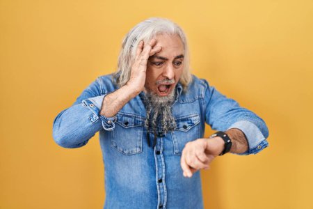 Téléchargez les photos : Homme du Moyen Âge avec les cheveux gris debout sur fond jaune regardant le temps de la montre inquiet, peur de se faire tard - en image libre de droit