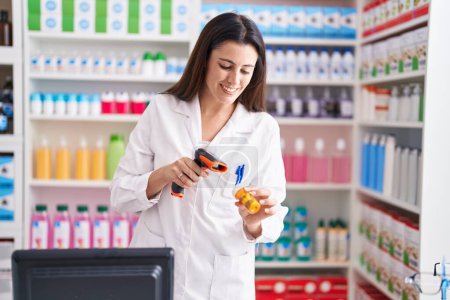 Foto de Joven hermosa mujer hispana farmacéutico escaneo píldoras botella en la farmacia - Imagen libre de derechos
