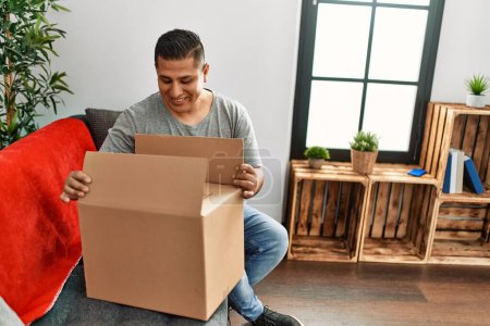 Photo for Young hispanic man smiling happy unboxing cardboard box sitting on the sofa at home. - Royalty Free Image
