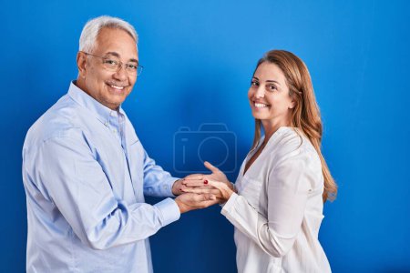 Foto de Mediana edad pareja hispana de pie sobre fondo azul señalando a un lado con las manos abiertas palmeras mostrando espacio de copia, presentando anuncio sonriente emocionado feliz - Imagen libre de derechos