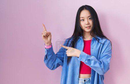 Foto de Young asian woman standing over pink background smiling and looking at the camera pointing with two hands and fingers to the side. - Imagen libre de derechos